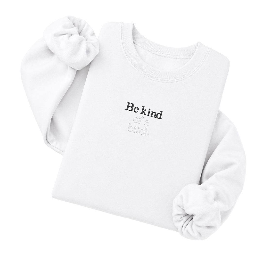 White crewneck sweatshirt embroidered with Be kind in black thread and of a bitch in tonal thread below