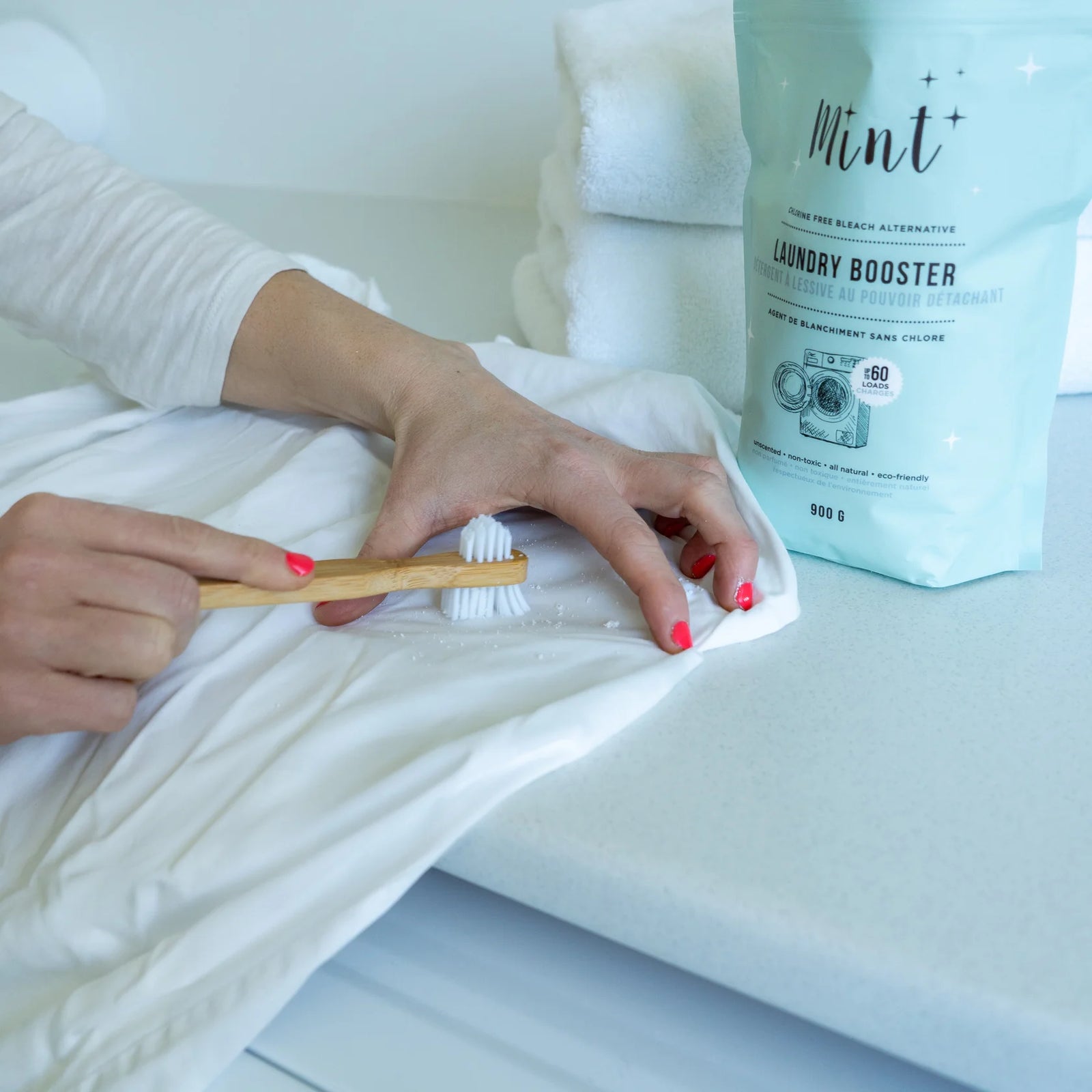 Person cleaning a white pillow with a wooden brush, next to a bag of Mint laundry booster.